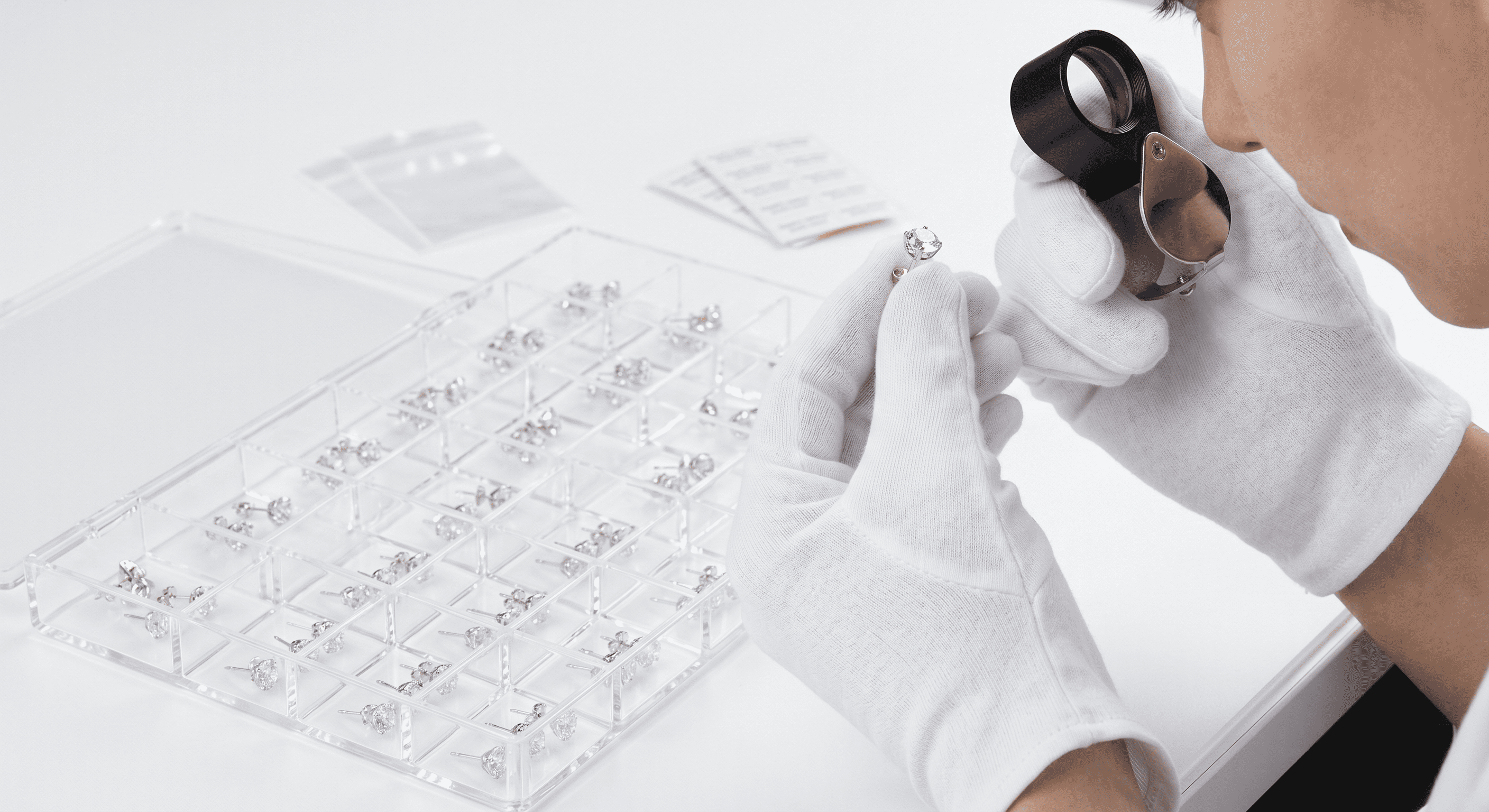 A quality control technician wearing immaculate white cotton gloves holds a silver stud earring up to a professional optical jeweler's loupe (1)