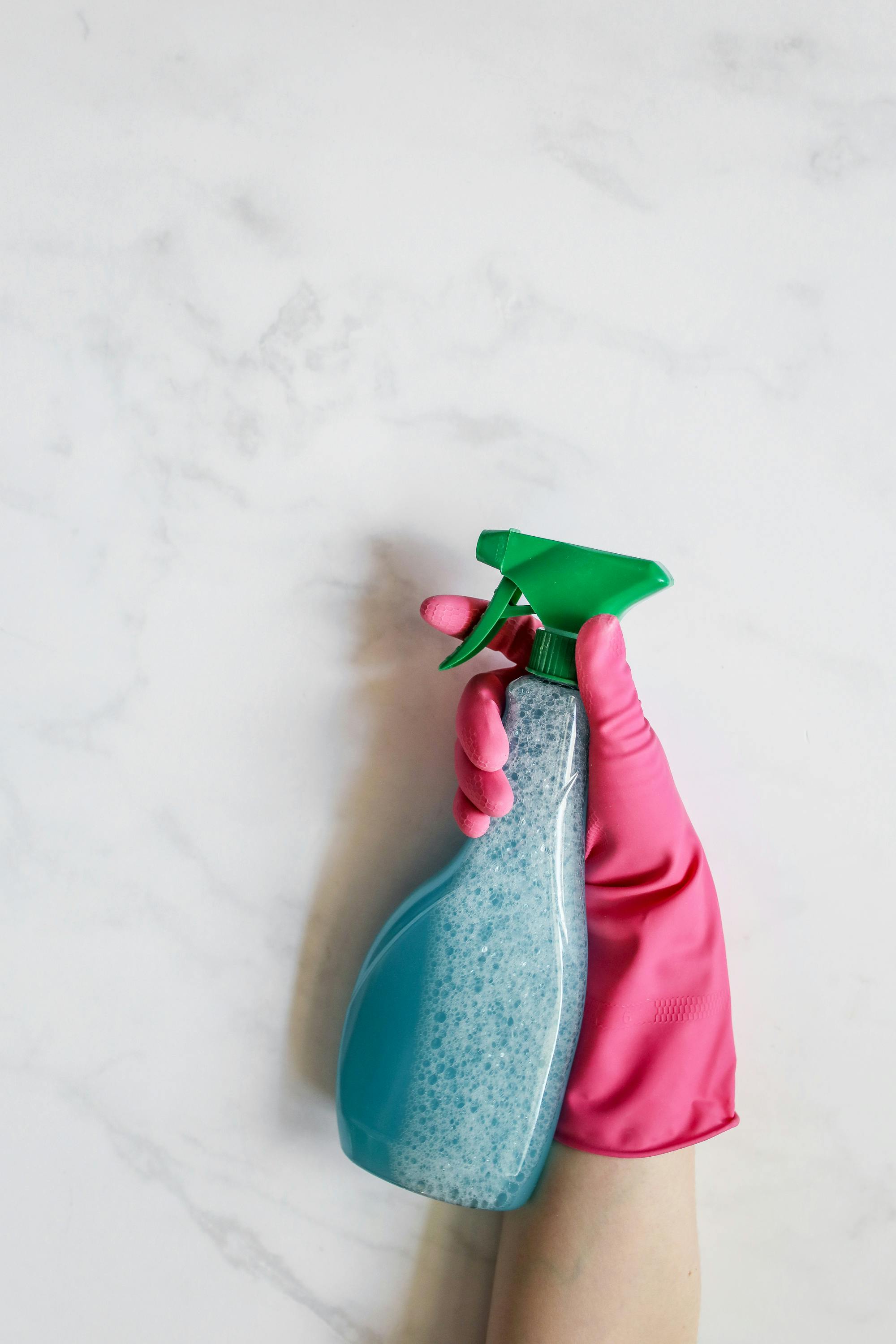 Imagen de primer plano de una mano con un guante rosa que sostiene una botella de spray sobre un fondo blanco. Imagen de primer plano de una mano con un guante rosa que sostiene una botella de spray sobre un fondo blanco.