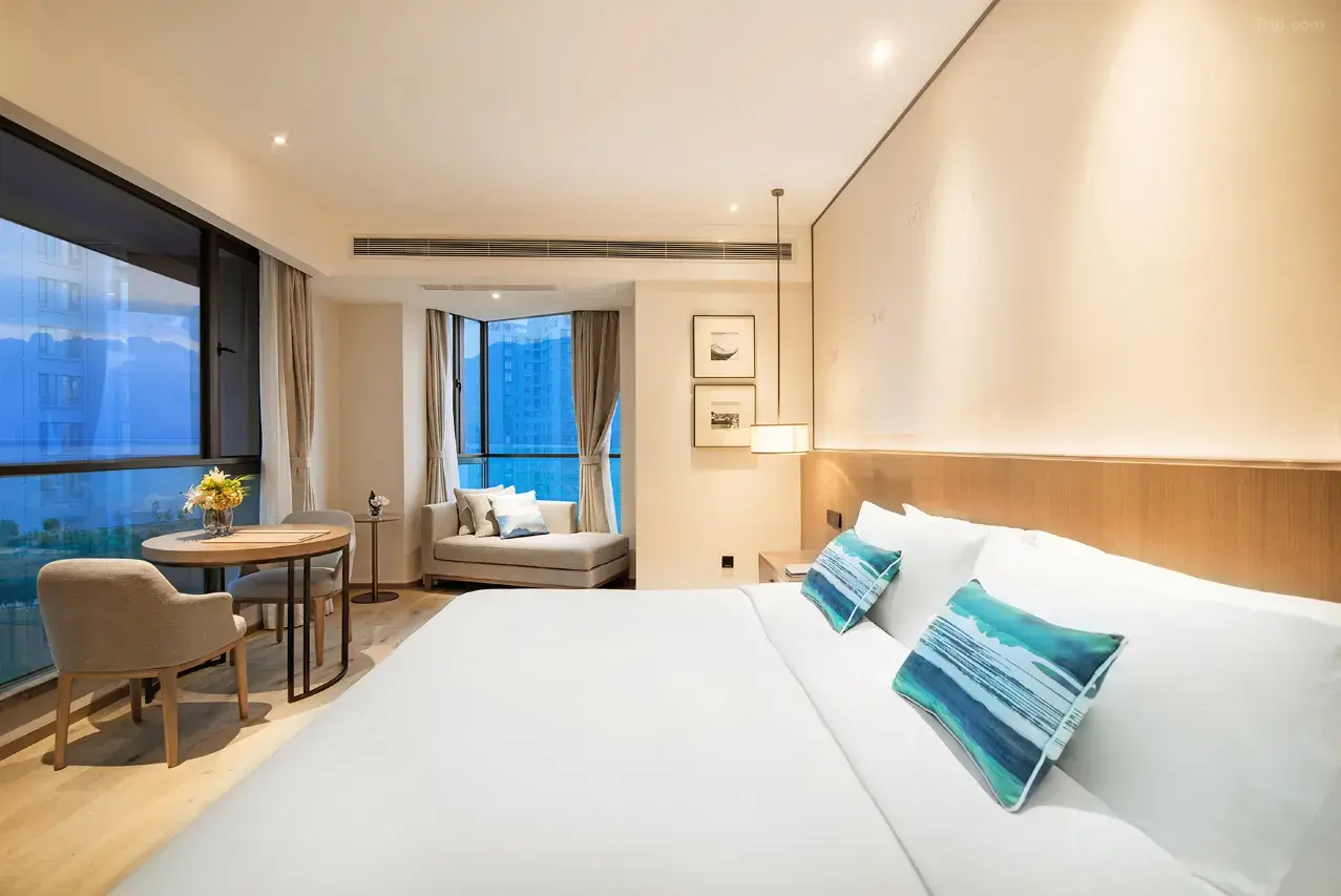 [主语种]Modern hotel room with large bed, sitting area, and city view from window.