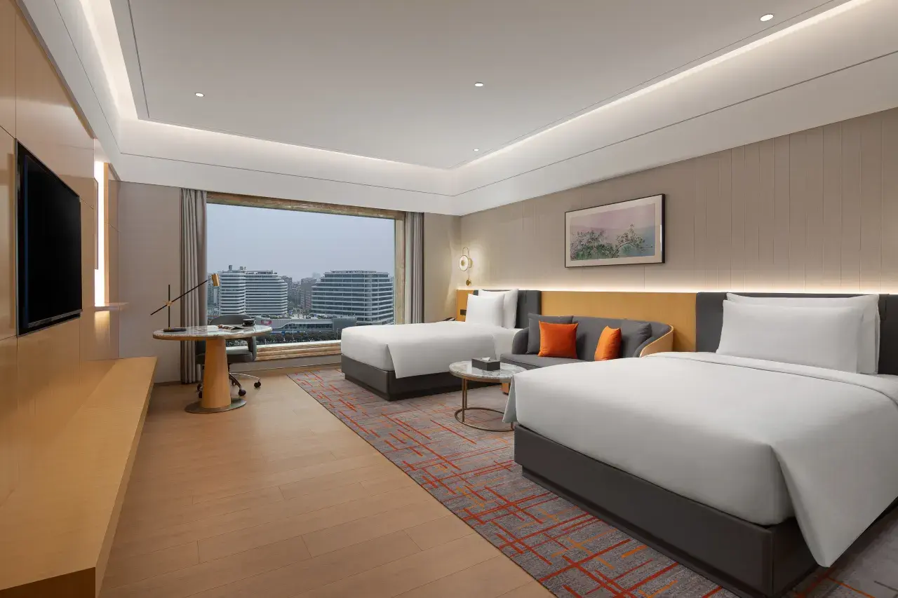 Modern hotel room with two beds, city view, sofa, and desk.