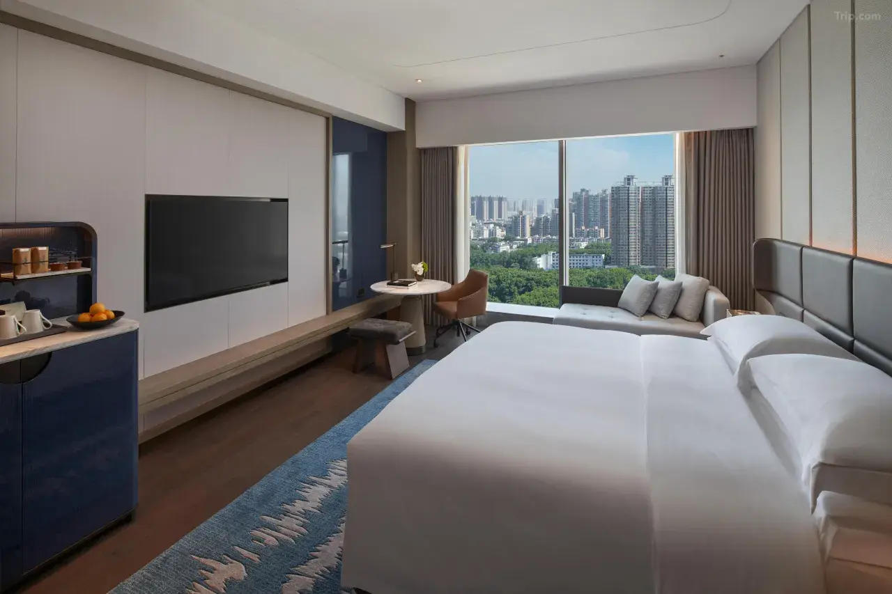 Modern hotel room with a large bed, TV, and floor-to-ceiling city view window.