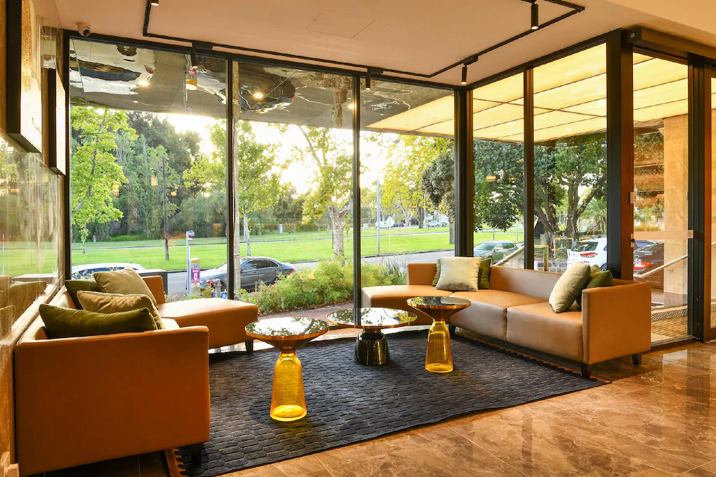 Lobby with brown sofas, green cushions, glass tables, and a view of trees and cars outside.