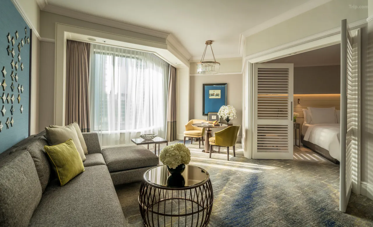 A modern luxury hotel suite with a gray sofa, dining area, and bedroom beyond.
