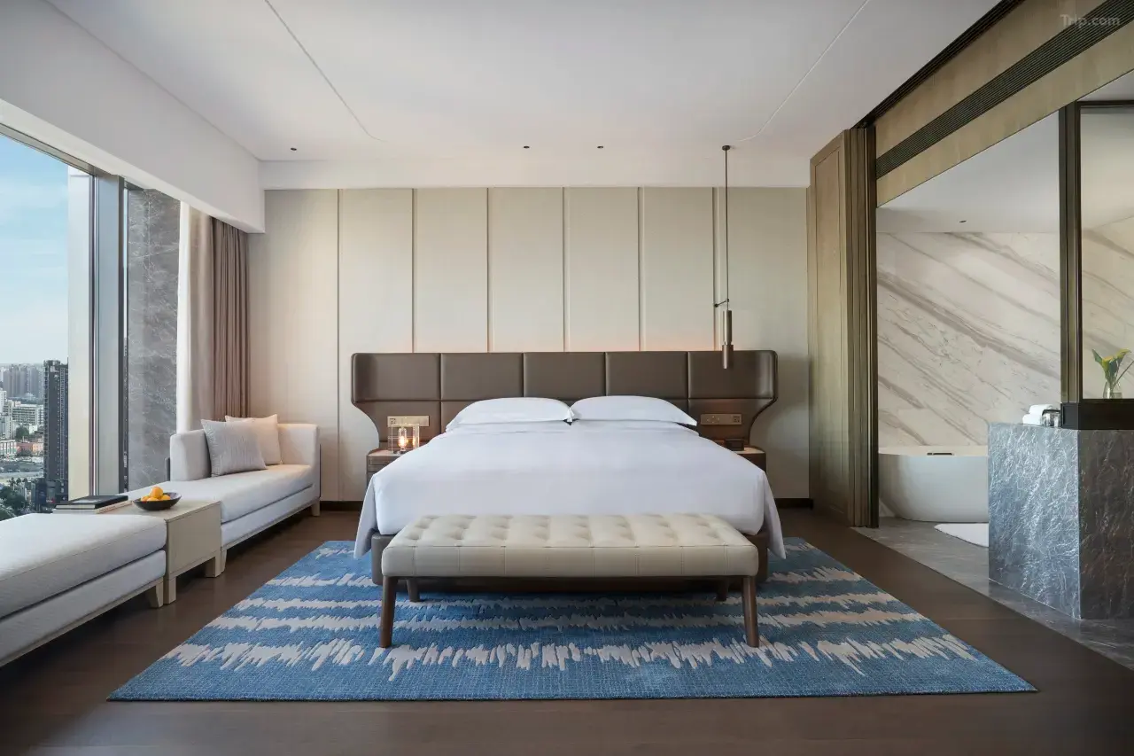 Modern luxury hotel suite with a large bed, blue rug, and city view.