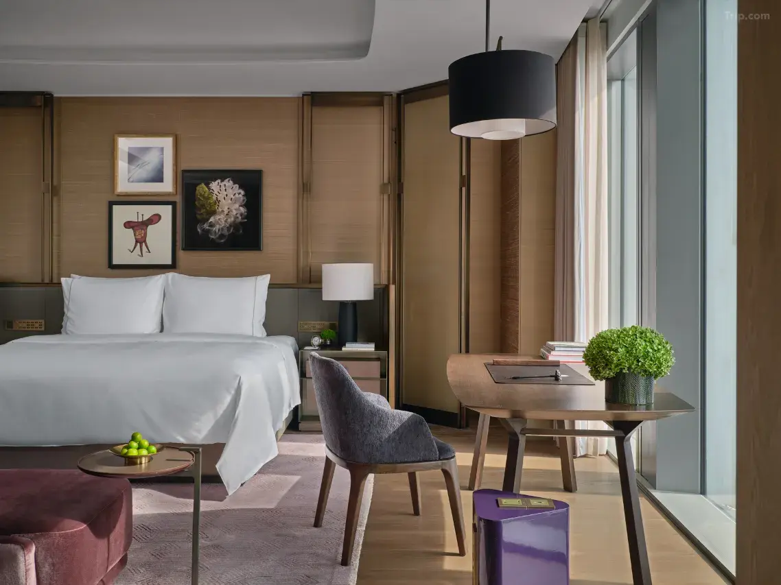 Modern hotel room with white bed, wooden desk, and large windows.