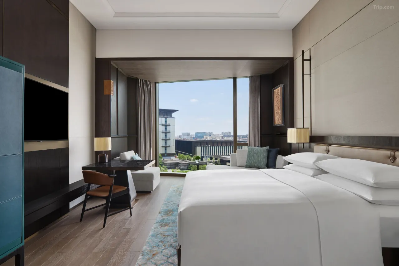 Modern hotel room with a large bed, desk, and city view through a large window.
