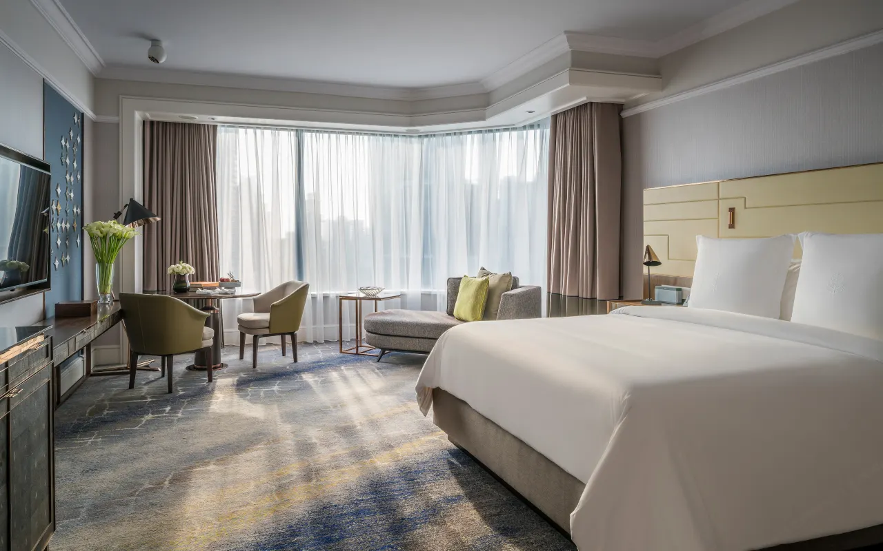Modern luxury hotel room with large windows, white bed, and seating area.