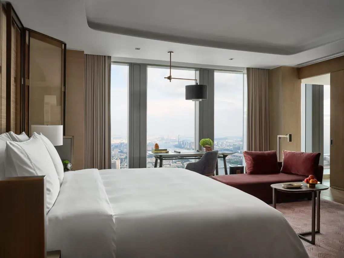 Modern hotel bedroom with a city view, desk, and red chaise lounge.
