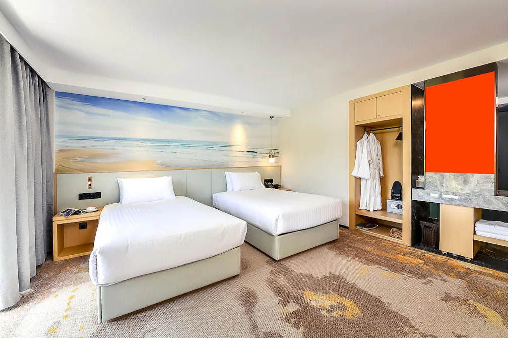 Twin beds in a hotel room with a beach mural, wardrobe, and modern decor.