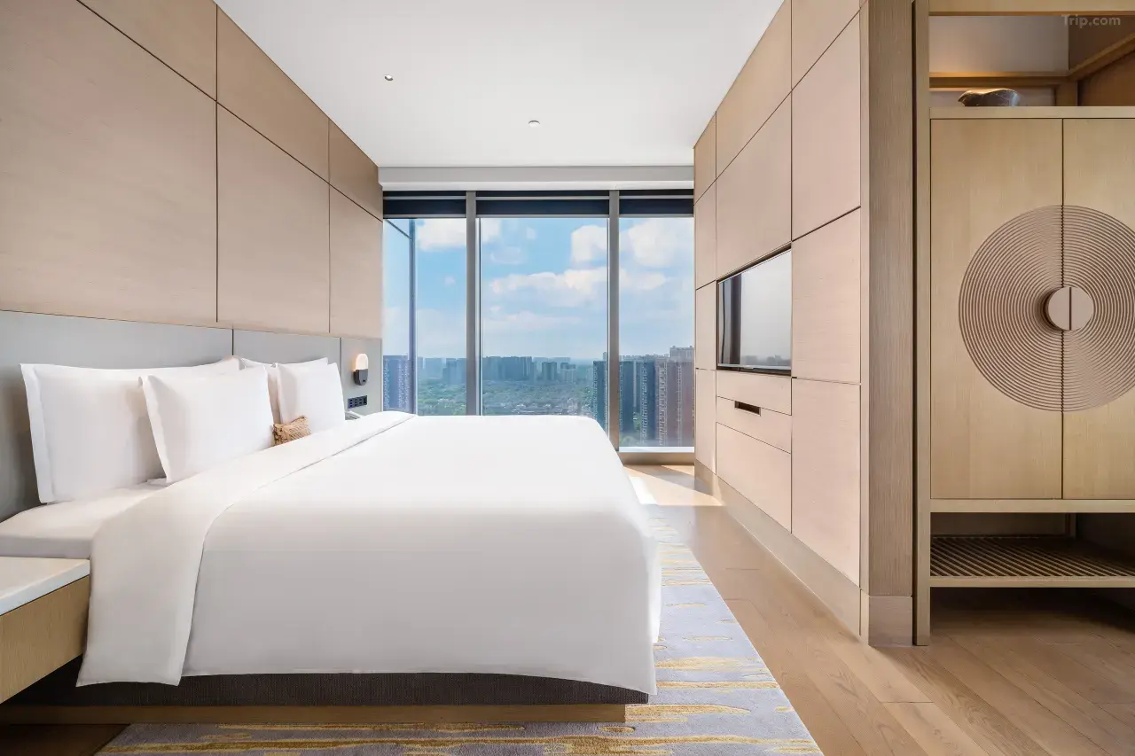Modern hotel room with white bed, wood paneling, and floor-to-ceiling city view.