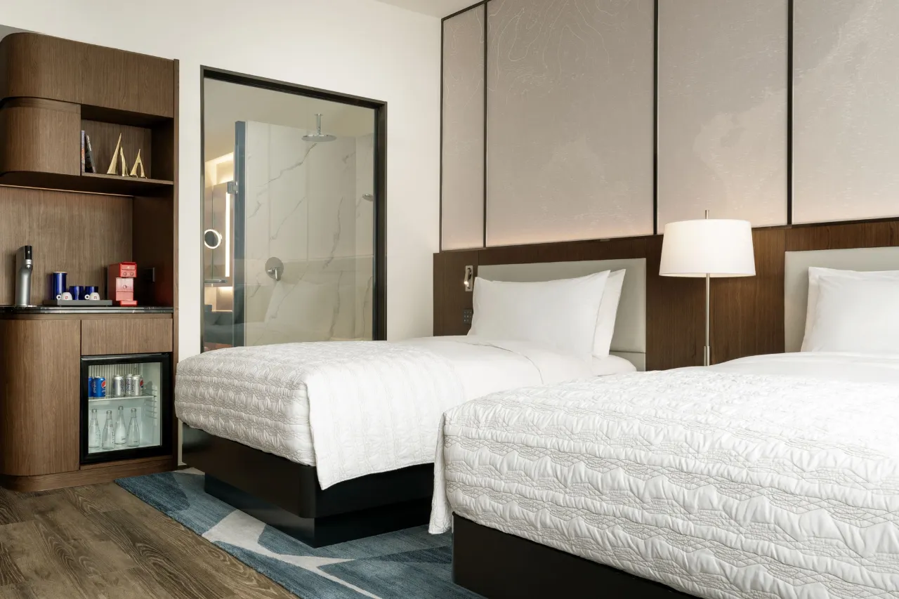 Modern hotel room with two twin beds, a mini-bar, and a glass shower.