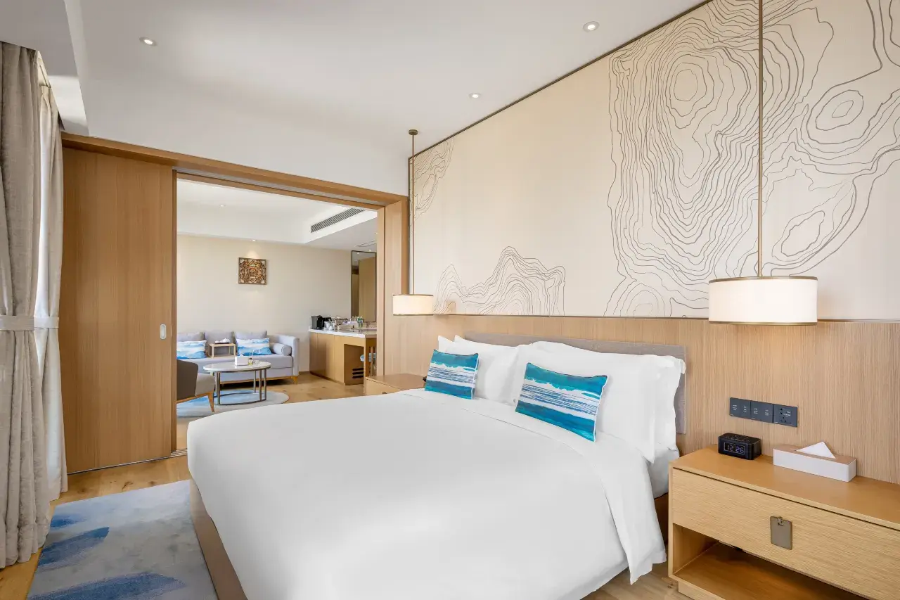 Bedroom with white bed, blue pillows, wood furniture, and a living area visible through an open door.