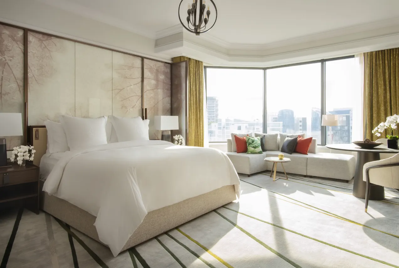 Modern hotel suite with a large bed, seating area, and floor-to-ceiling windows.
