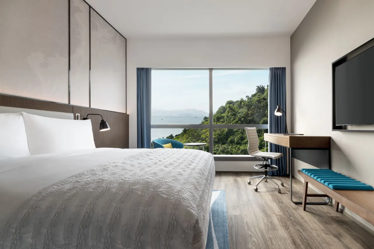 Modern hotel room with a large window overlooking the ocean and lush greenery.