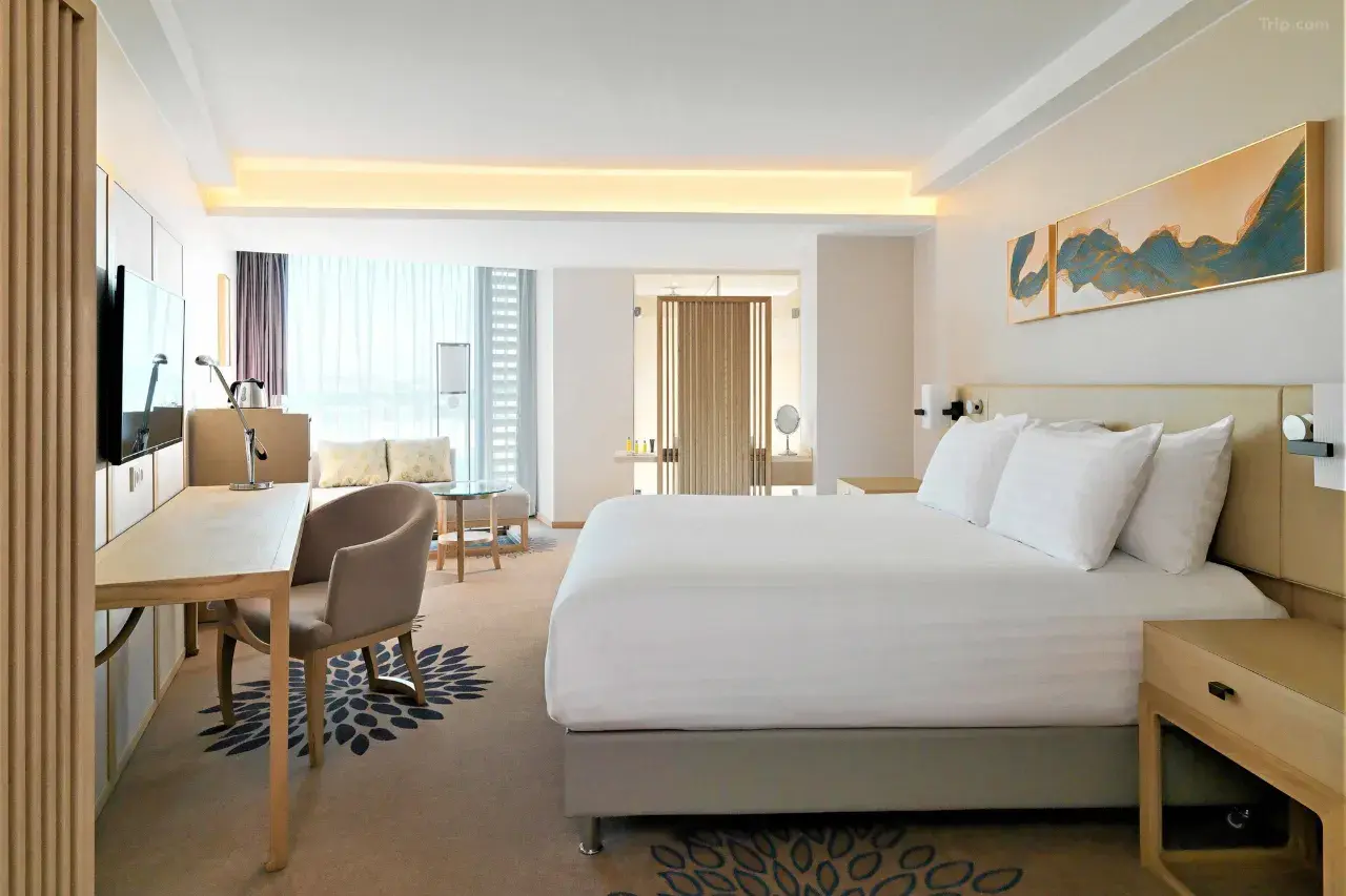 Modern hotel room with white bed, desk, and large window.