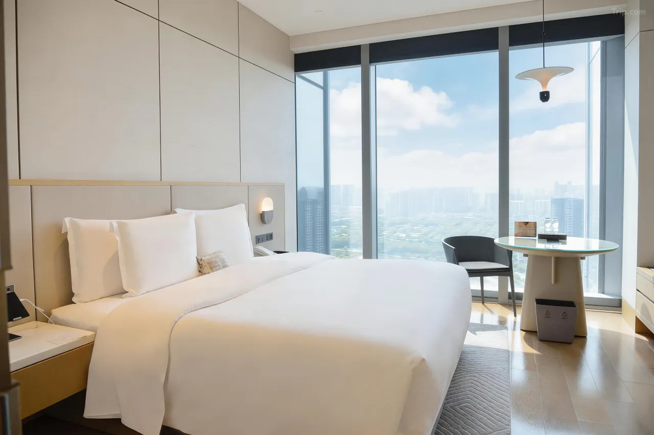 Modern hotel room with a white bed and floor-to-ceiling city view windows.