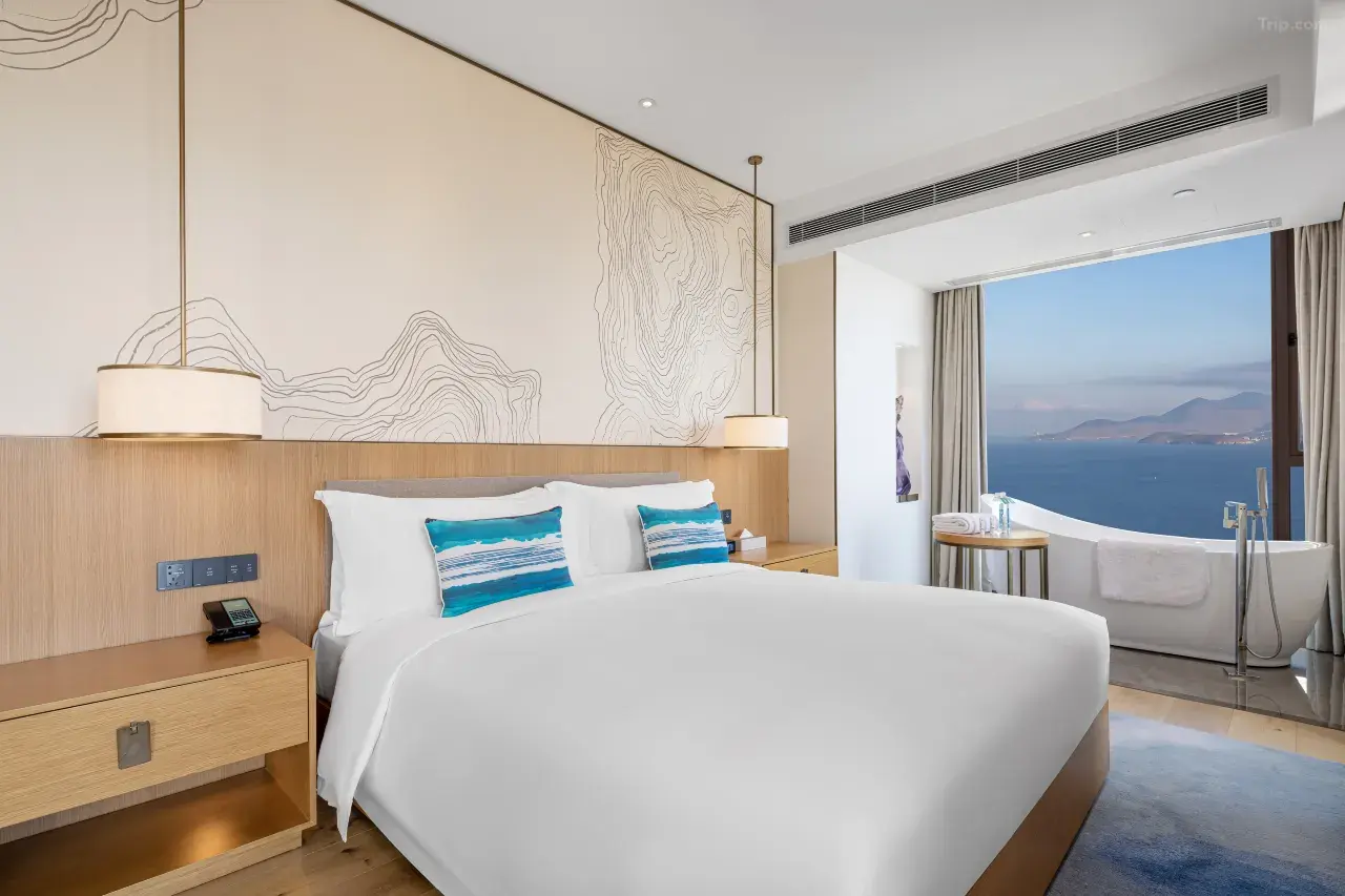 Modern hotel bedroom with white bed, blue pillows, and a bathtub overlooking the ocean.
