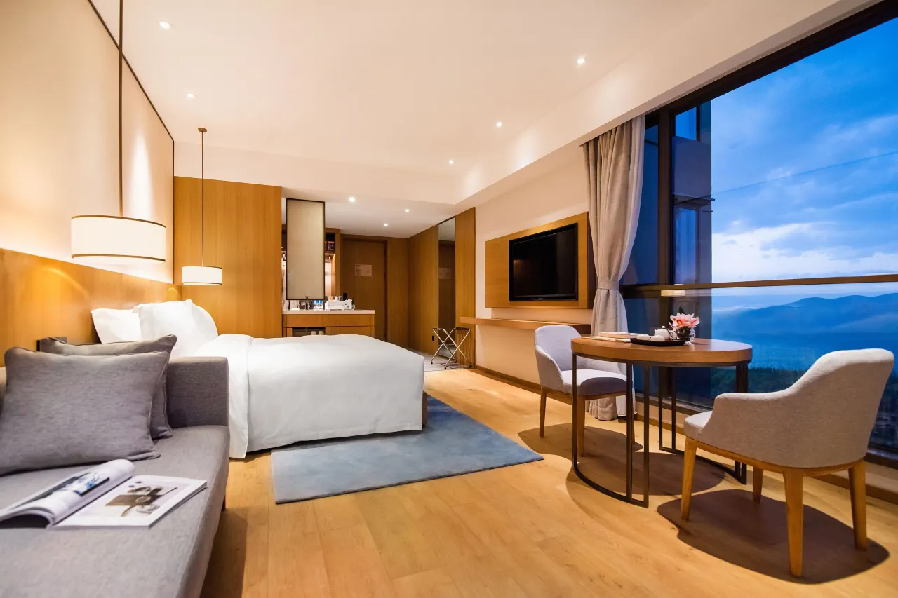 Modern hotel room with a large window overlooking mountains under a cloudy sky.