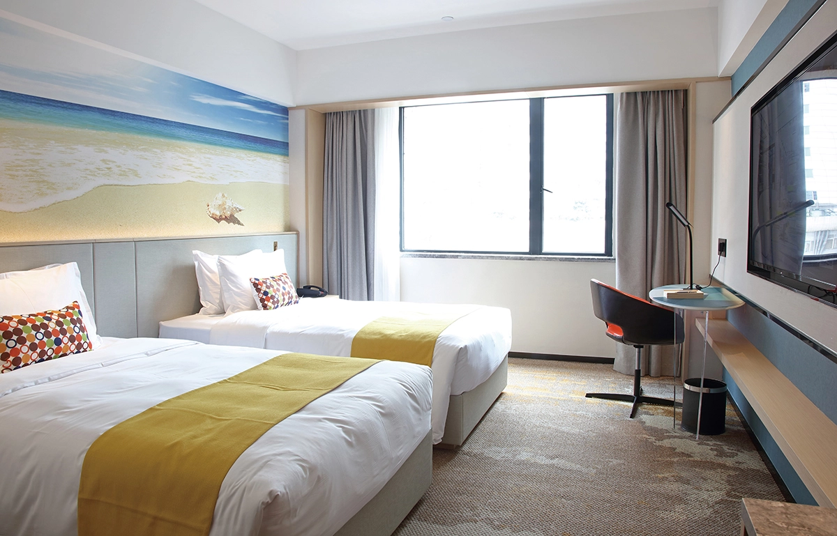 Twin beds in a modern hotel room with beach mural, large window, and TV.