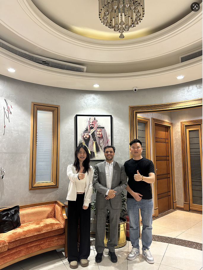 Three people pose in front of a portrait of Saudi Arabian rulers in an ornate room.
