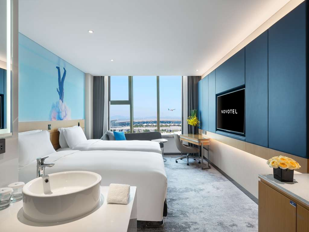 Novotel hotel room interior with two beds, sink, and airport view.