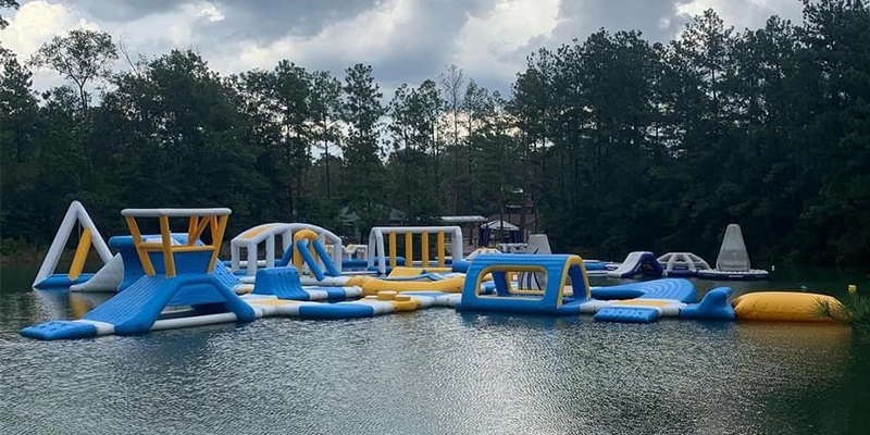 An image of an inflatable water park with a variety of obstacles and slides. The water park is located in a lake surrounded by trees.