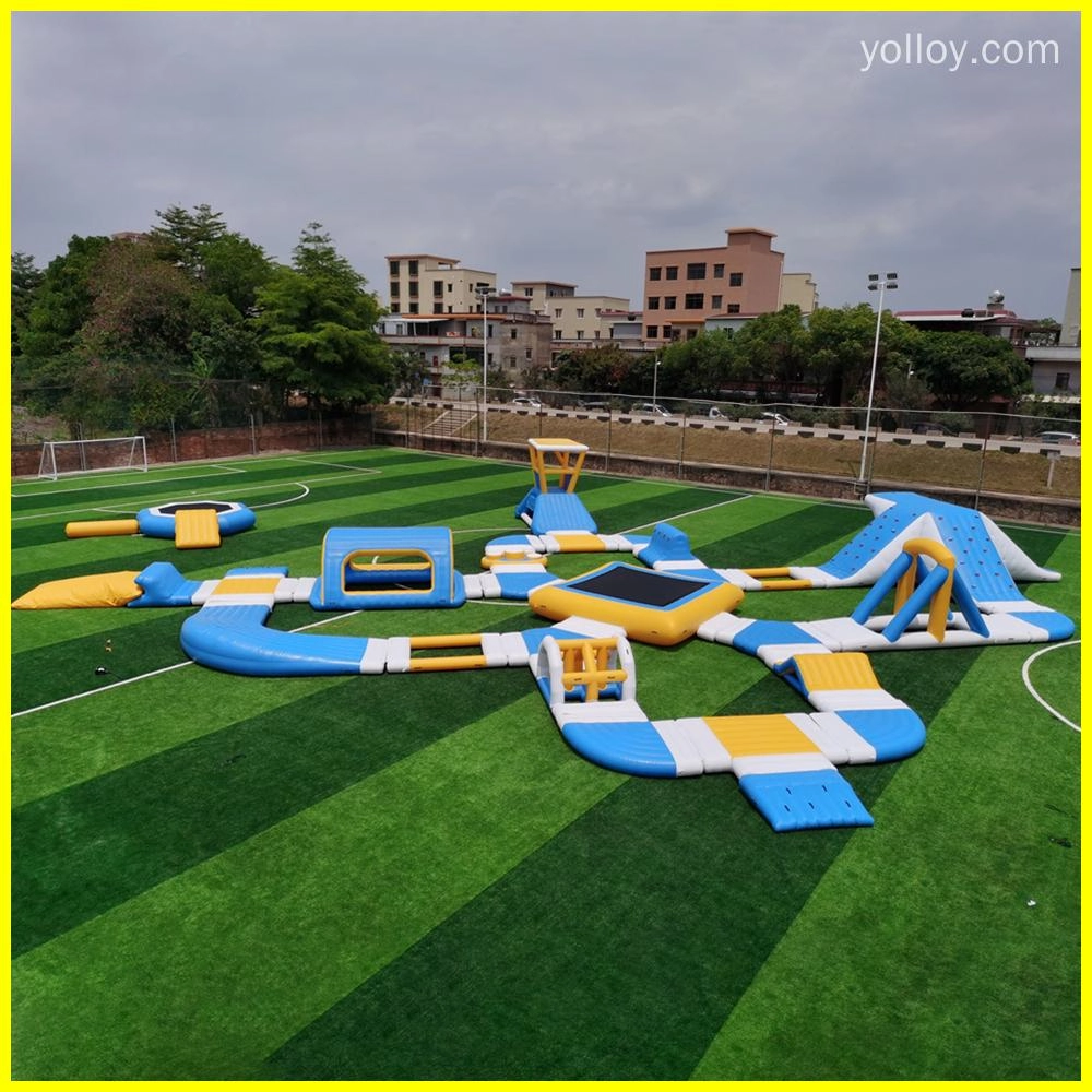 Imagen de un parque acuático inflable con diversas características, como toboganes, trampolines y pistas de obstáculos. Está instalado en un campo verde con árboles y edificios de fondo.