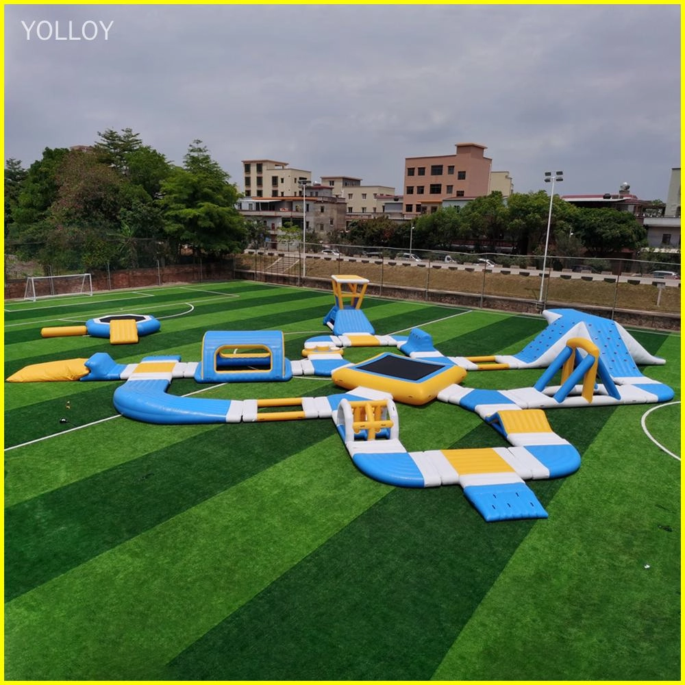 An image of an inflatable water park with various features such as slides, trampolines, and obstacle courses. It is set up on a green field with trees and buildings in the background.