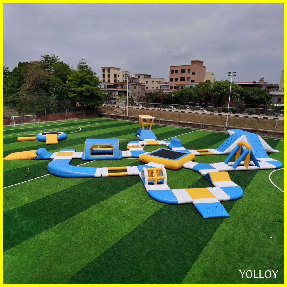 An image of an inflatable water park with various features such as slides, trampolines, and obstacle courses. It is set up on a green field with trees and buildings in the background.