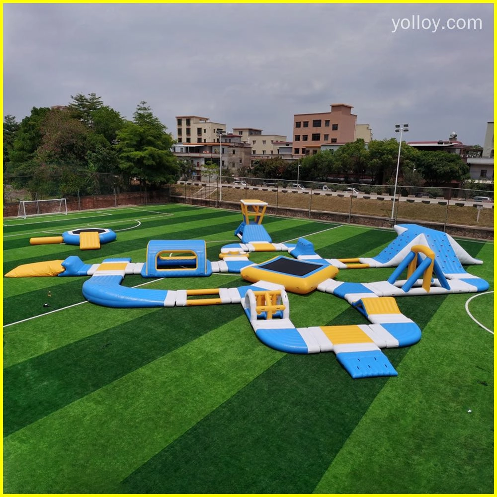 Uma imagem de um parque aquático inflável com vários recursos, como escorregadores, trampolins e pistas de obstáculos. Ele é montado em um campo verde com árvores e prédios ao fundo.