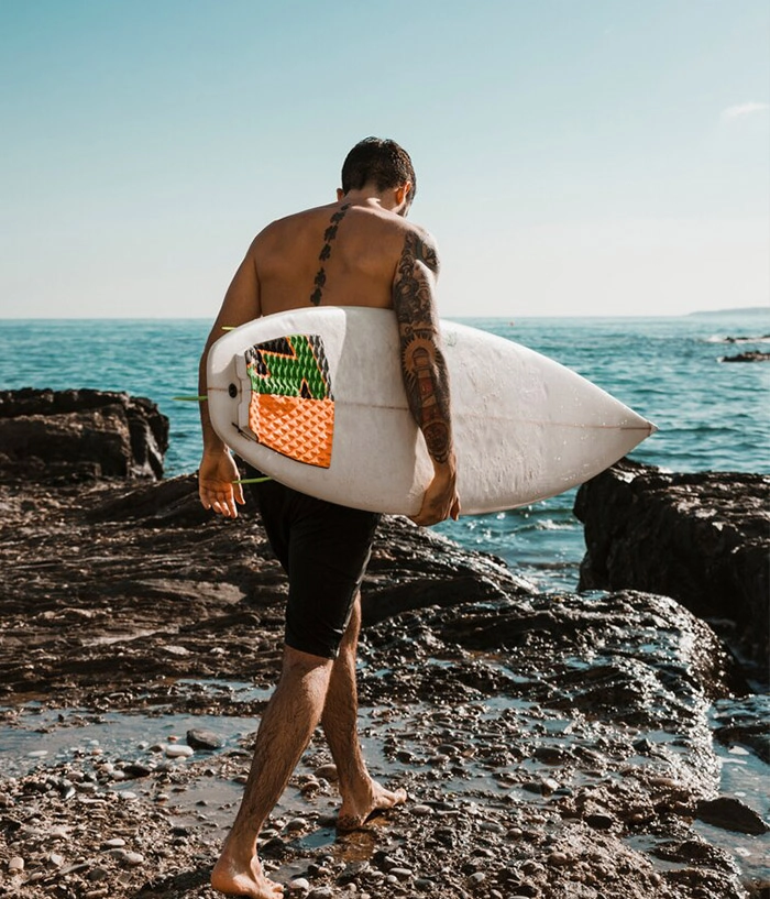 Un homme avec des tatouages ​​​​sur le dos et le bras marche sur la plage en portant une planche de surf.