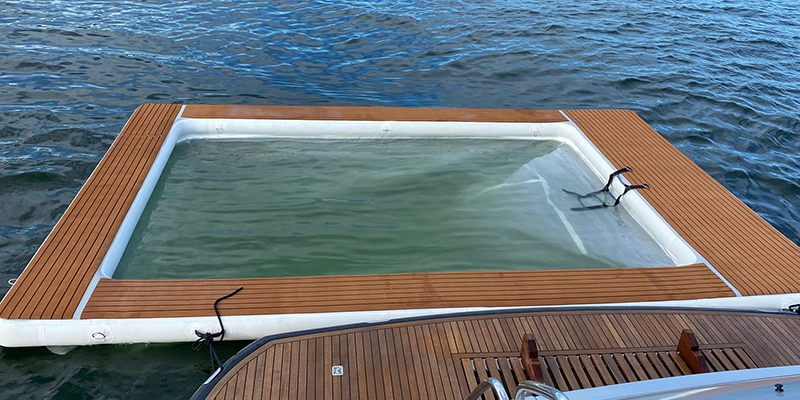 An inflatable pool floats on the water next to a boat. The pool is made of white PVC and has a wooden deck.