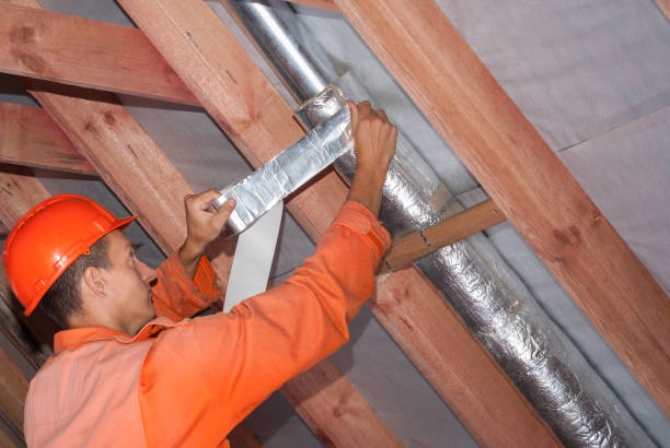 A construction worker in an orange jumpsuit and hard hat uses metallic tape to seal insulation around a duct in an attic. - FUNAS