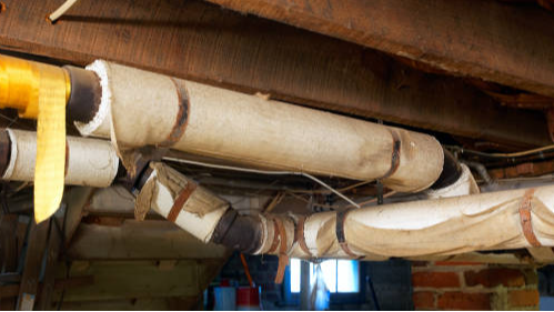 Old, insulated pipes running along a wooden basement ceiling.