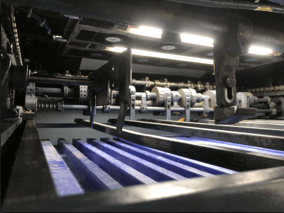 Close-up view of the interior mechanism of an industrial machine, showing intricate metal parts, a series of blue ribbed components, and bright lights illuminating the machinery.