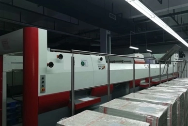 Wtjd  The image shows a large, automated bookbinding machine in a factory. The machine is surrounded by stacks of paper and books.