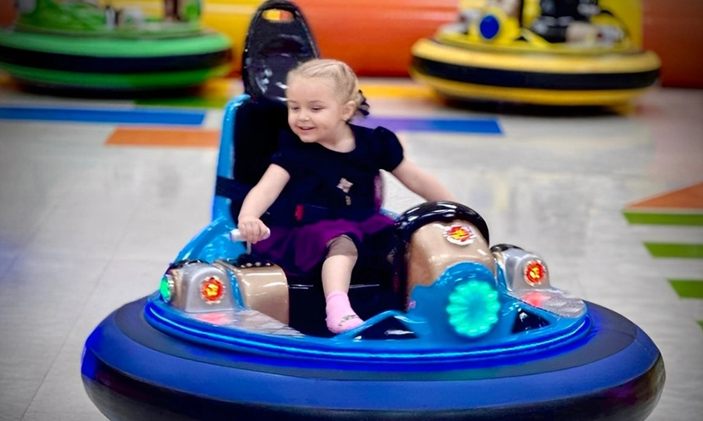 A young girl with pigtails smiles as she rides a blue bumper car - H.Q.Amusement