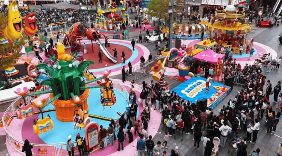 Parque de diversões ao ar livre movimentado com a montanha-russa SUNHONG Larva Blooms-16P no canto inferior esquerdo, o Carrossel Larva no canto superior direito e grandes figuras do personagem Larva ao fundo - SUNHONG