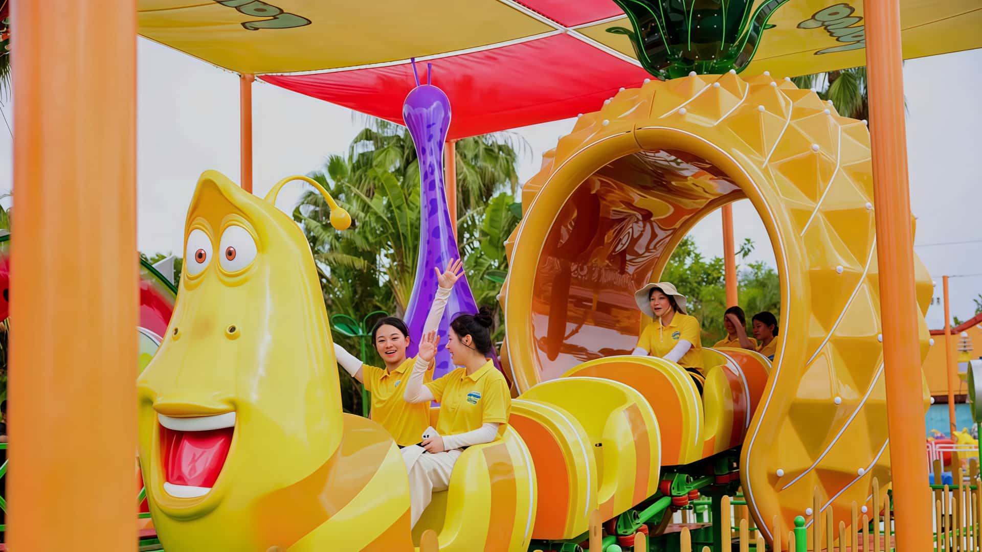 Five female passengers laughing and enjoying the Larva Fruitland mini roller coaster ride, passing through a pineapple-shaped tunnel at Larva Adventure Theme Park in Seaside Bay. Theme park solutions providing thrilling experiences.