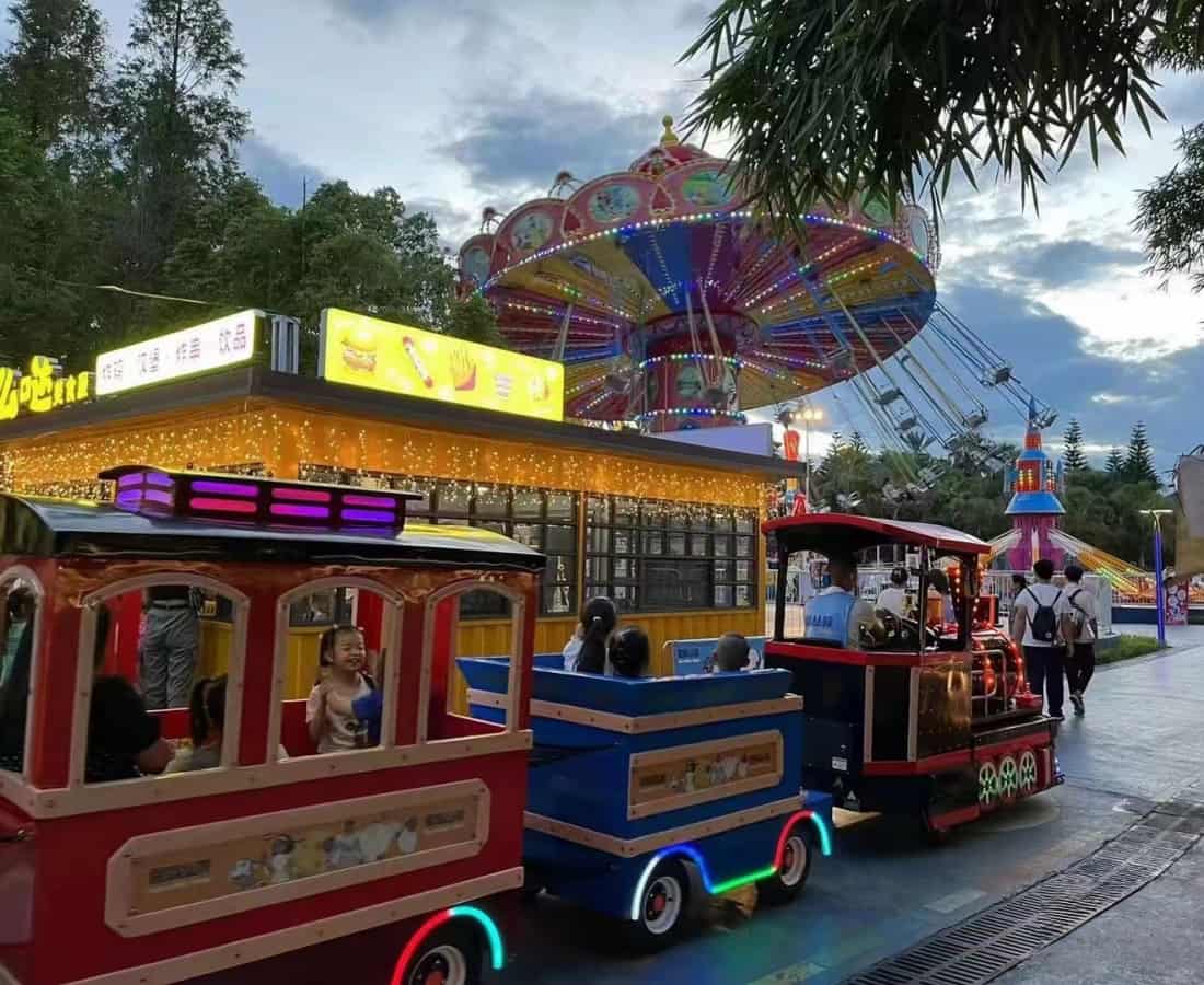 أطفال يركبون قطارًا مصغرًا في مدينة ملاهي، مع ألعاب التأرجح وأكشاك الطعام في الخلفية، لعرض حلول قطارات الملاهي من SUNHONG لمناطق الترفيه العائلي - SUNHONG