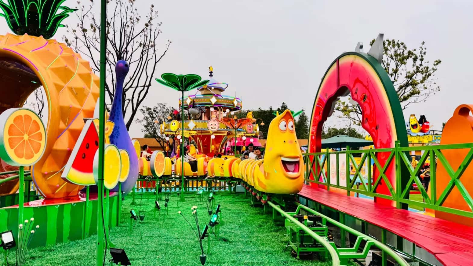 Larva Fruitland Mini Roller Coaster in Operation at Larva Theme Park The Larva Fruitland Mini Roller Coaster running at Larva Theme Park, with a yellow-themed train head coming toward the camera, and the Larva Carousel in the background.