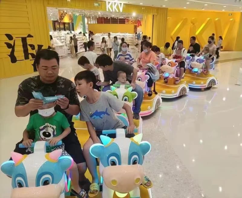 أشخاص يركبون لعبة أطفال ملونة على شكل بقرة من إنتاج شركة SUNHONG في مدينة ملاهي داخلية مضاءة بشكل ساطع، لعرض حلول ألعاب الملاهي المناسبة للعائلات في أماكن الترفيه الداخلية - SUNHONG