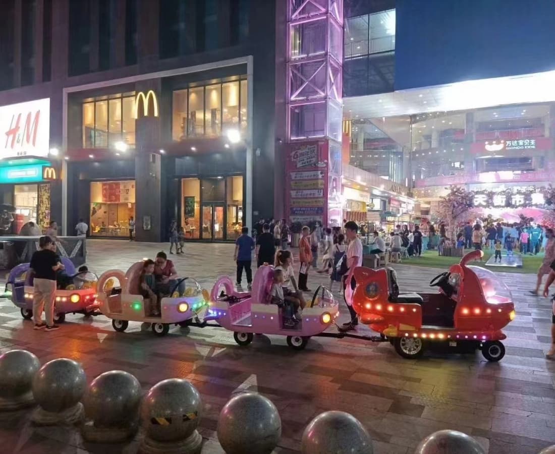 Trem elétrico colorido sem trilhos para crianças operando à noite em frente à entrada movimentada de um shopping. - SUNHONG