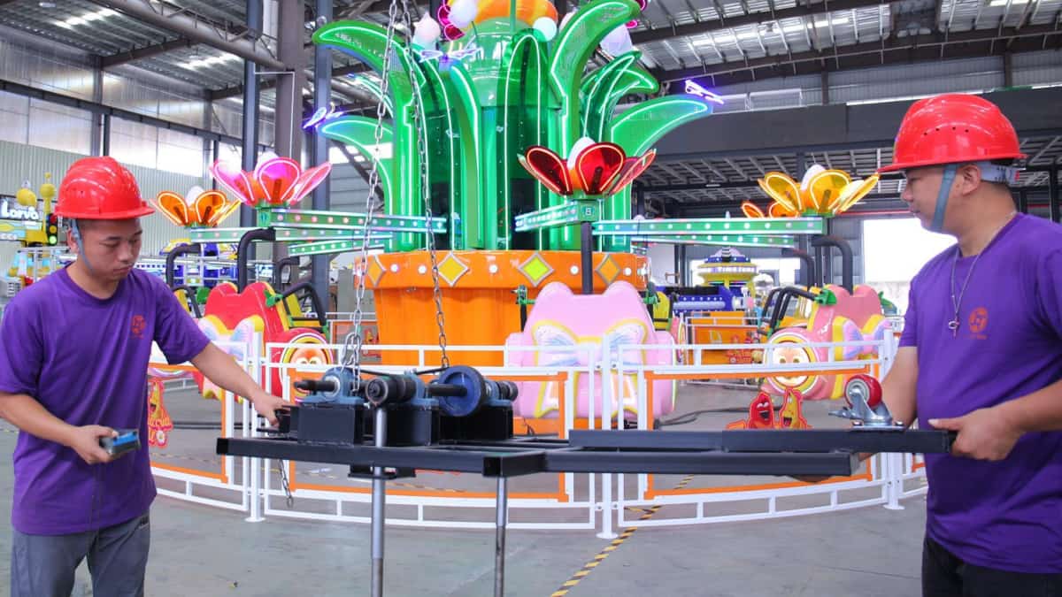 Two employees at SUNHONG Amusement Rides Manufacturer using a lift to move parts in front of Larva Blooms, preparing for installation. - SUNHONG