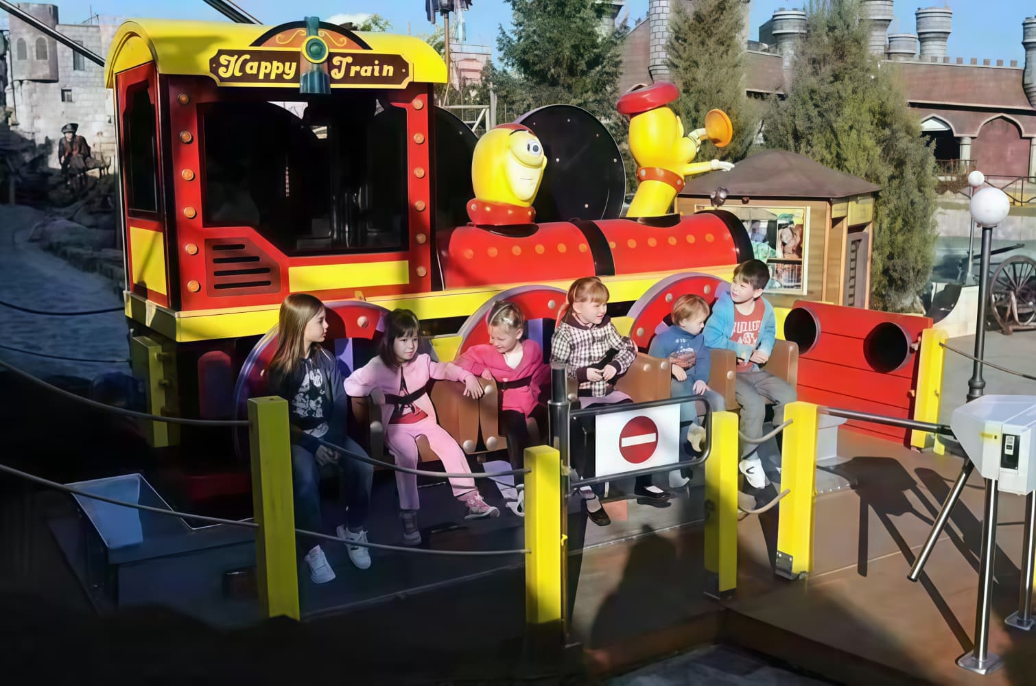 Six children sitting on a colorful Happy Train amusement park ride on a sunny day.