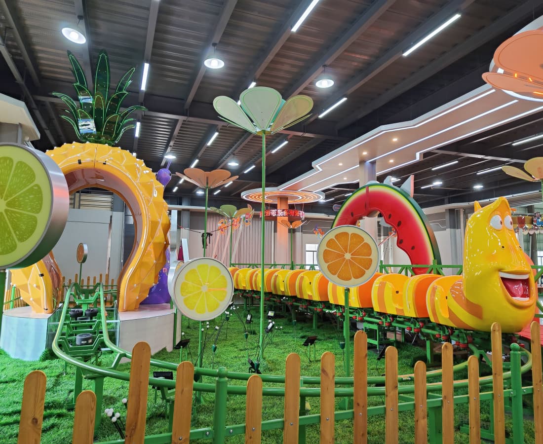 Larva Fruitland mini roller coaster on display at SUNHONG showroom, featuring Yellow character, seats, green track, and a pineapple tunnel. - SUNHONG