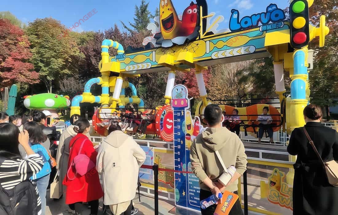 Larva_Swing_Theme_Park_Queue_2025.jpg Larva swing ride in operation at a theme park, with a long line of visitors waiting to experience the thrilling fair ride.