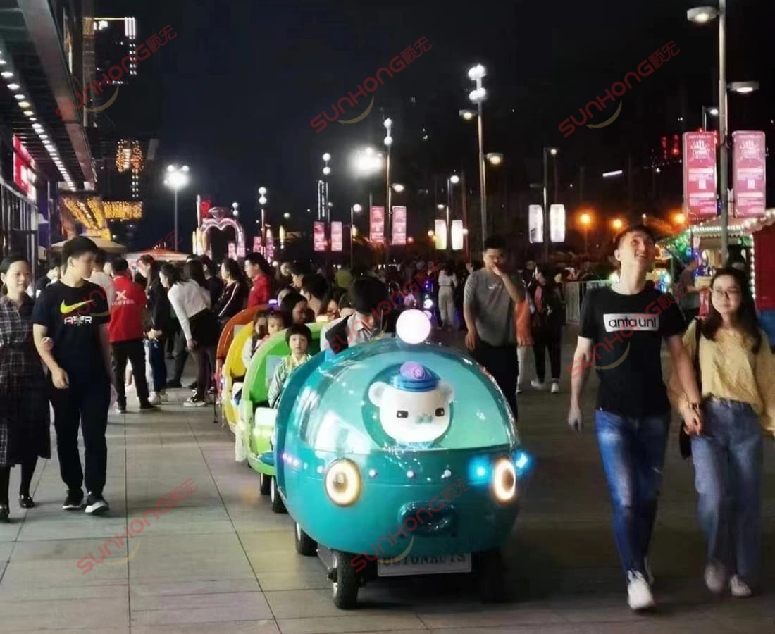 octonauts-13p-trem-sem-trilhos-cidade-rua - SUNHONG Vista frontal dos vagões de trem sem trilhos Octonauts 13P circulando em uma movimentada rua de pedestres à noite, atraindo famílias. - SUNHONG