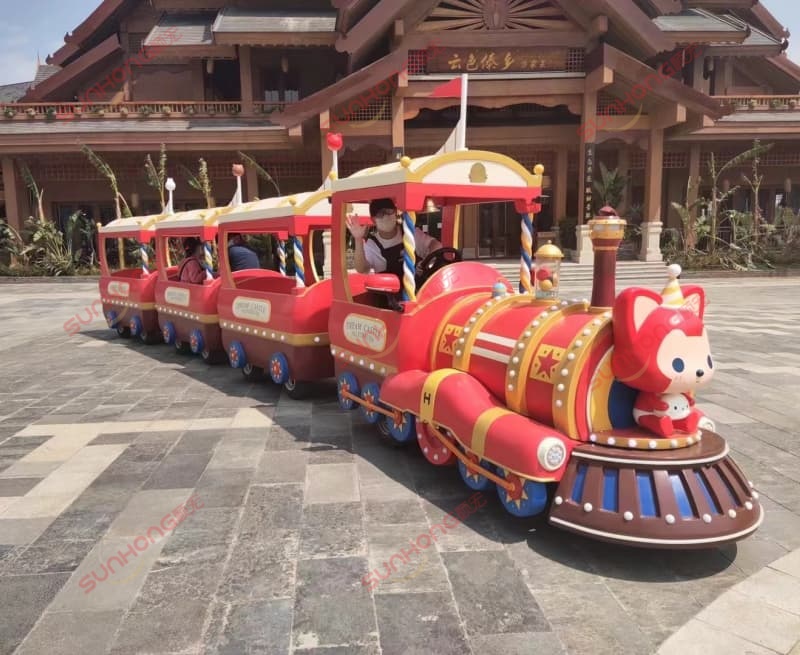 sunhong-amusement-park-train-ride-red-fox-cartoon-kids-train Red SUNHONG children's amusement park train ride with a cartoon fox character, carrying passengers in front of a building in a theme park environment