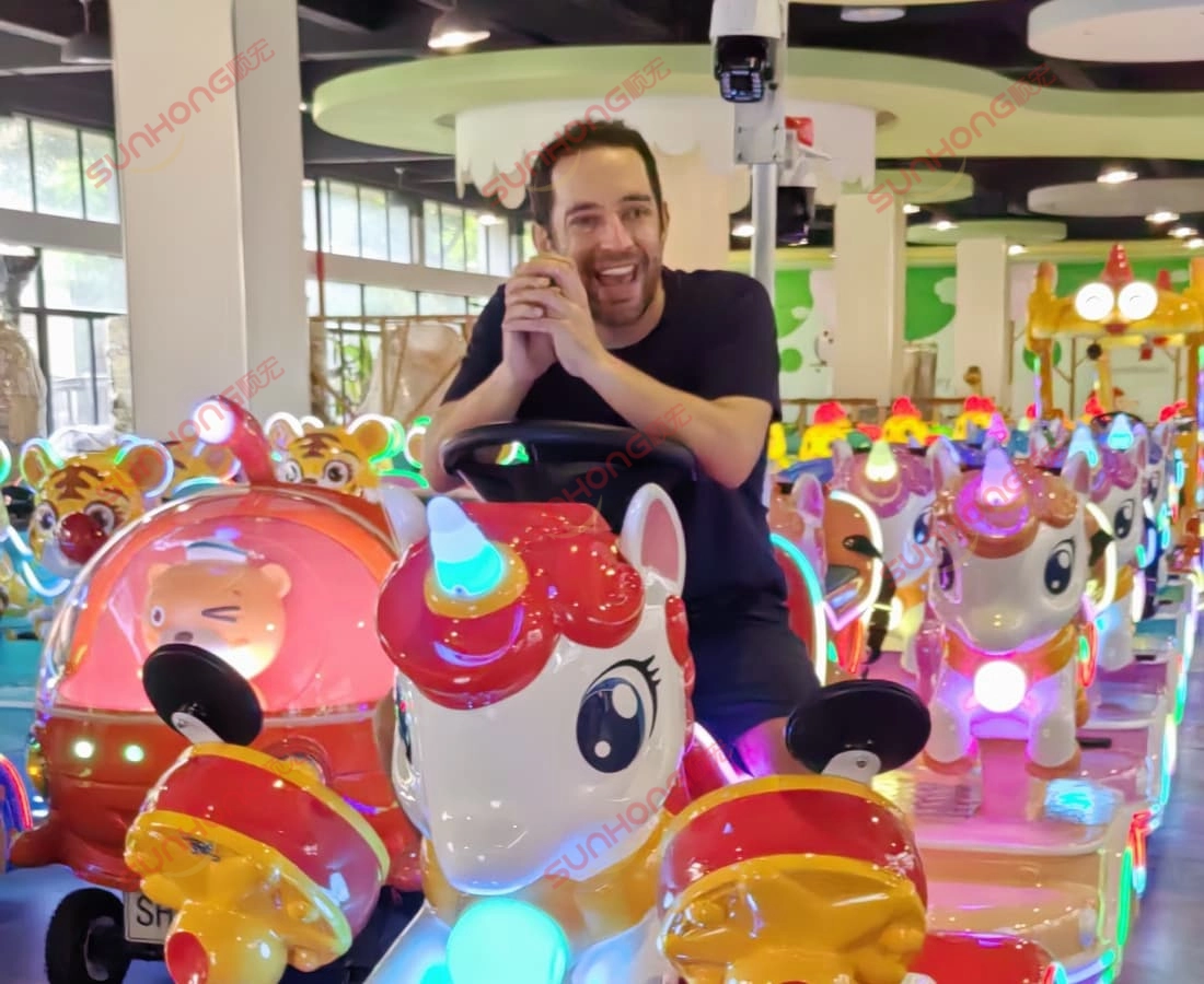 unicorn-trackless-train-ride-happy-dewasa-indoor - SUNHONG Lelaki dewasa tersenyum menaiki kereta api kanak-kanak unicorn di taman hiburan tertutup berwarna-warni dengan lampu terang. - SUNHONG